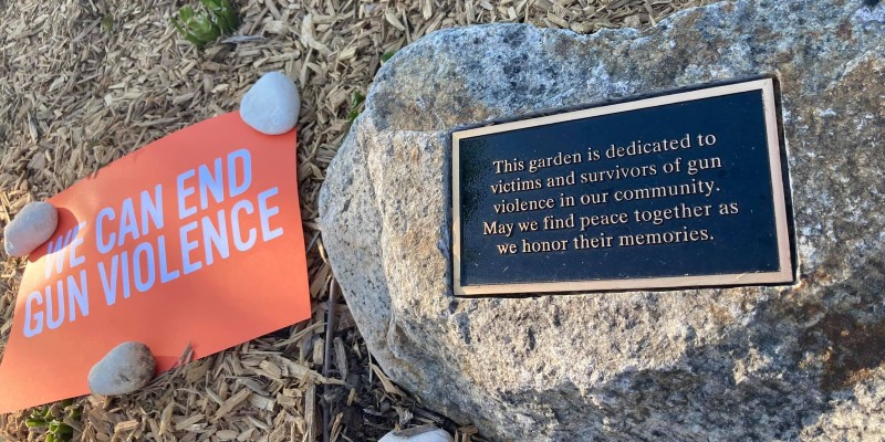 Photo of a rock with a commmorative plate acknowledging lives lost to gun violence. On the left of the image is an orange sign that says 