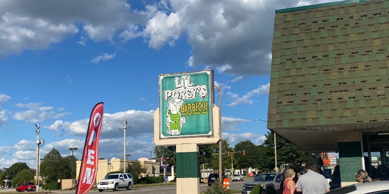 Photo of Lil Porgy's sign against a blue sky with puffy white and gray clouds. The rood of the restaurant is visible on the right. A red, vertical OPEN sign is on the left. Photo by Julie McClure. 