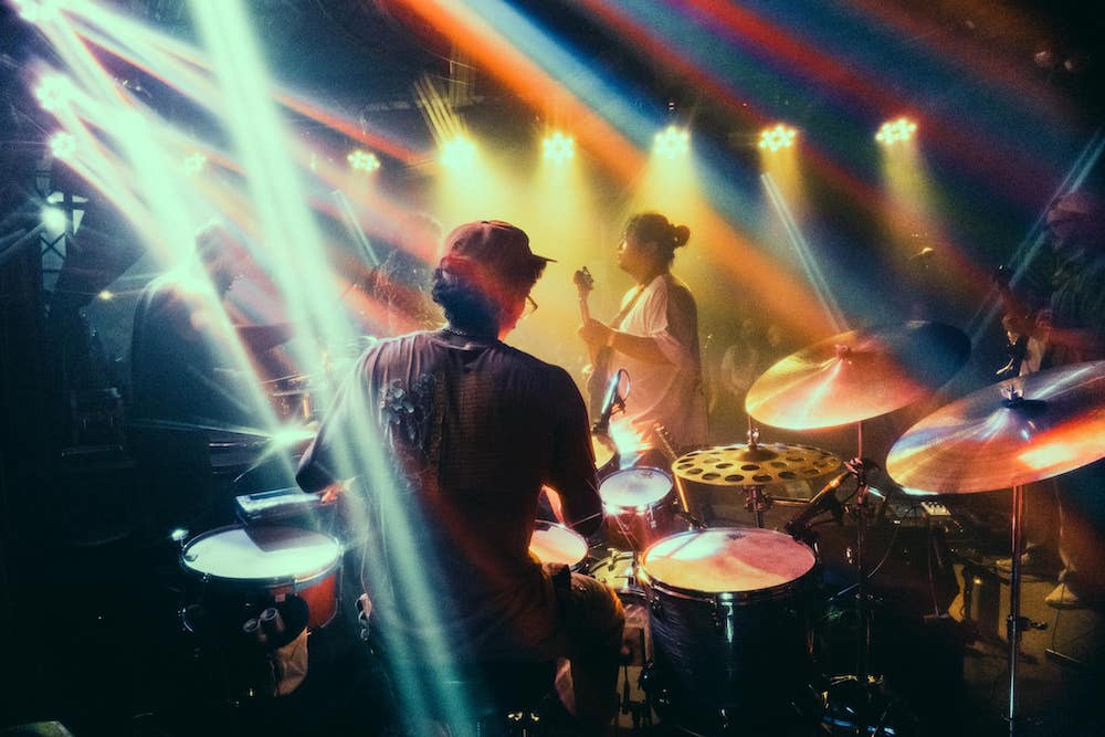 A view of a band on stage from behind the drummer. The drummer has a baseball cap on, and is sitting behind a drum set. In the foreground is a guitarist. There are beams of stage lights streaming across the stage. Photo from Zoofunkyou's Facebook page.