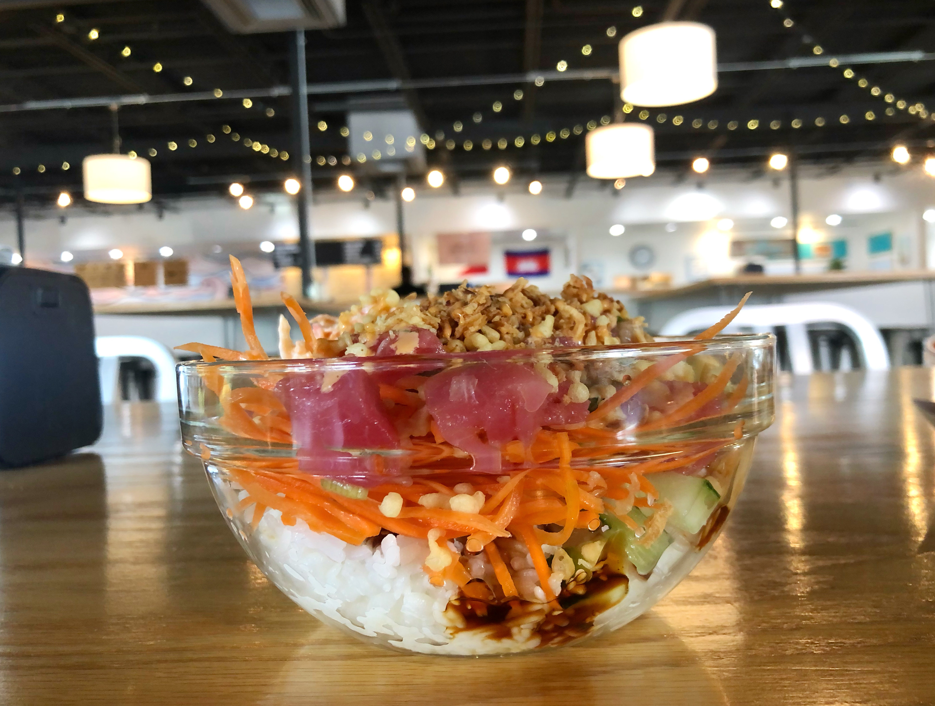 On a warm wooden table, there is a glass bowl of tuna poke with dark sauce dripping down white rice and tempura crunch on top. In the background, there are blurred string lights and blurred flags and signs of Broadway Food Hall restaurants. Photo by Alyssa Buckley.