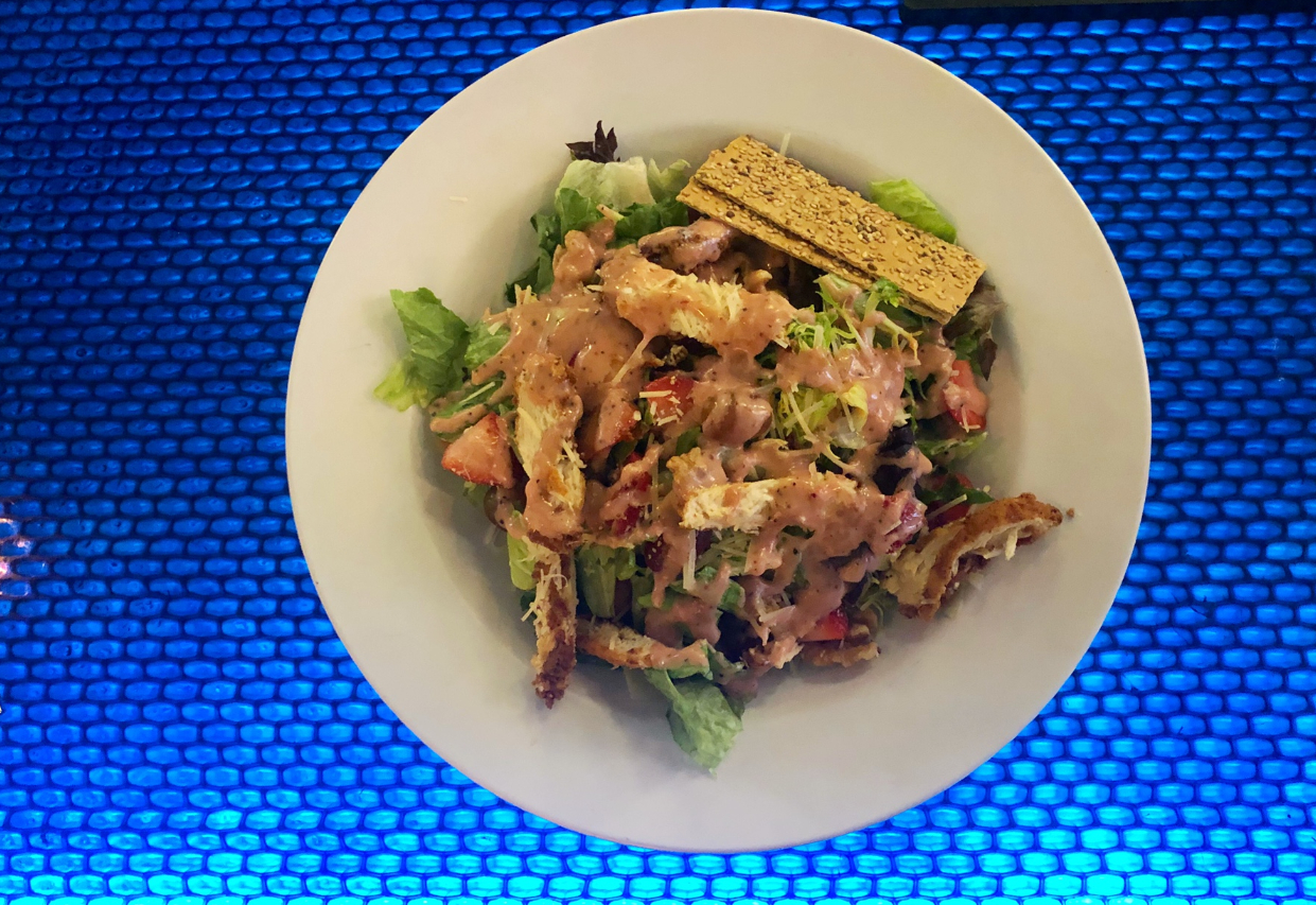 On a blue-lit bar, there is a white circular plate of salad with a pink poppyseed dressing, fried chicken pieces, and fruit. Photo by Alyssa Buckley.