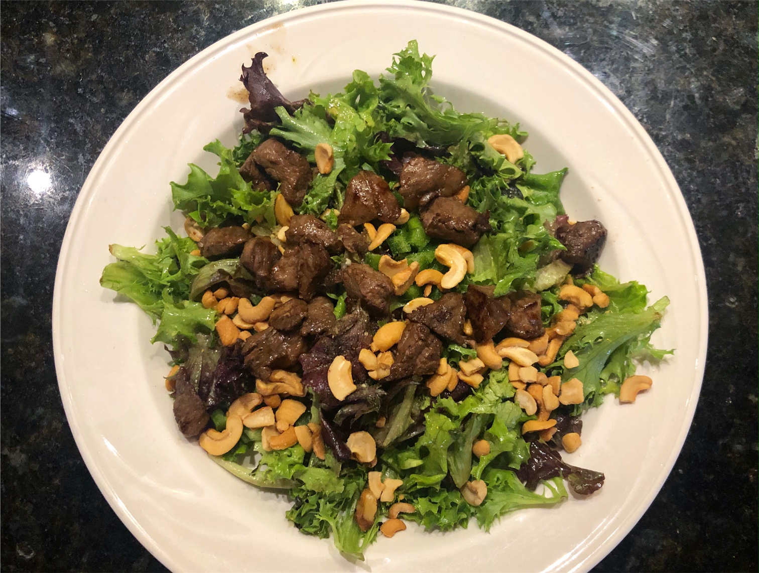On a white plate, there is a steak salad with lots of steak chunks, cashews, and greens. Photo by Alyssa Buckley.