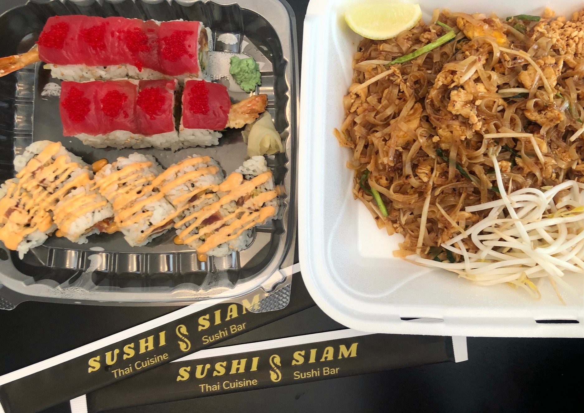 On a black table, there is a takeout order of sushi and pad Thai with two unopened chopsticks in black and white paper with the restaurant's name printed on it. Photo by Alyssa Buckley.