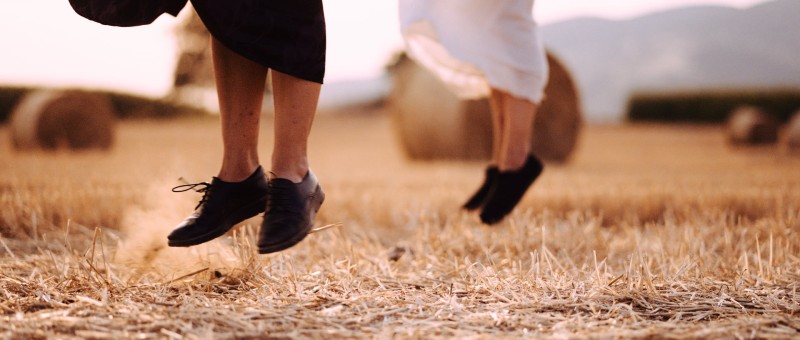 A close up of two people's feet mid-jump. They both have black shoes, and one is wearing a white dress and the other a black dress. They are in a field with bales of hay in the background. Photo from Flatlands Dance Film Festival Facebook page. 