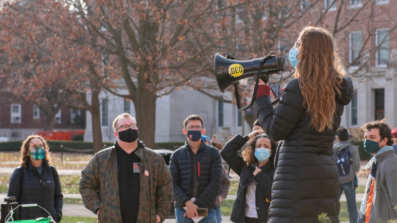 A woman with long brown hair and a black winter coat is holding a blue megaphone up. There are people wearing masks facing her. They are standing on the U of I quad. Photo by J. Sidney Malone.