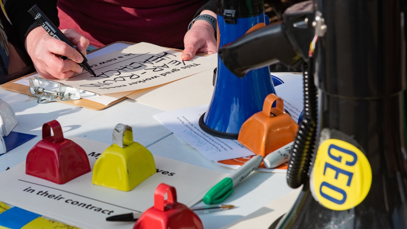 A close up of a hand holding a black marker. The person is writing YEAR-ROUND HEALTHCARE on a sign. There are colorful bells and a blue megaphone sitting on the table. Photo by J. Sidney Malone.
