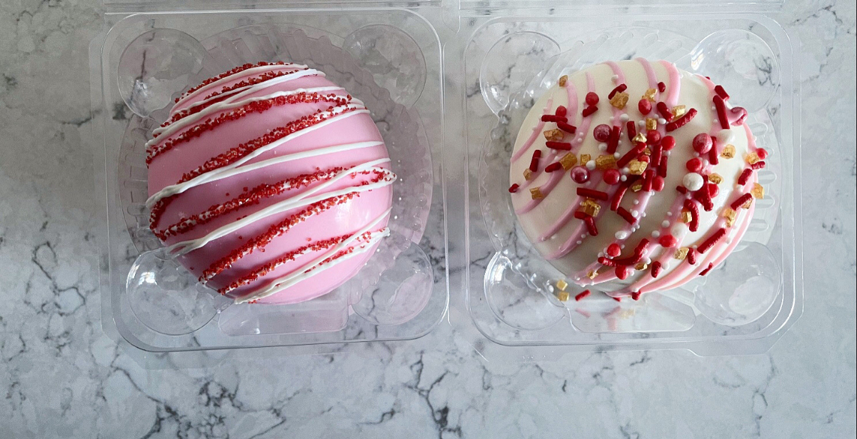 On a marble counter, there are two cocoa bombs in clear clamshell containers. The one on the left is pink with white chocolate drizzle and red sprinkles, and the one on the right is a white sphere with red and white sprinkles. Photo by Alyssa Buckley.