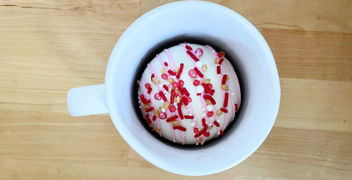 On a butcher block counter, there is a white mug with a white chocolate sphere of chocolate with red and white sprinkles. Photo by Alyssa Buckley.