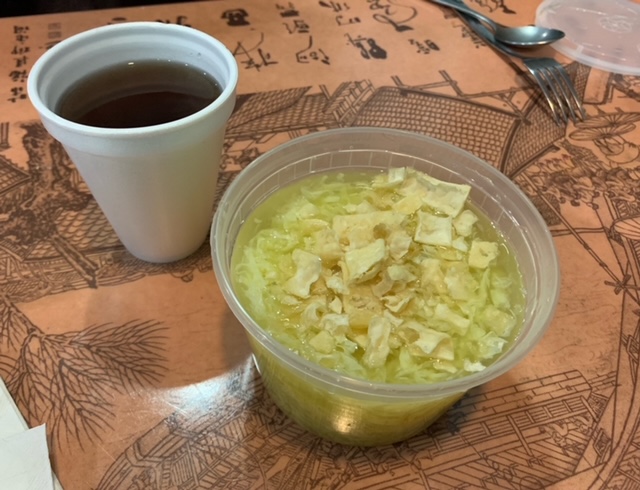 On a table, there is a plastic bowl of egg drop soup beside hot tea in a white styrofoam cup. Photo by Carl Busch.