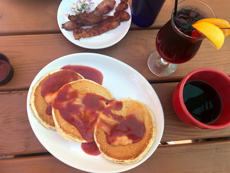 On a brown slatted table, there is an oval white plate with three yellow pancakes and a slather of red syrup. To the right, there is a coffee in a red mug, and a pink cocktail in a glass. To the top in the center, there is a small plate of diced red onions and two strips of cooked bacon. Photo by Alyssa Buckley.