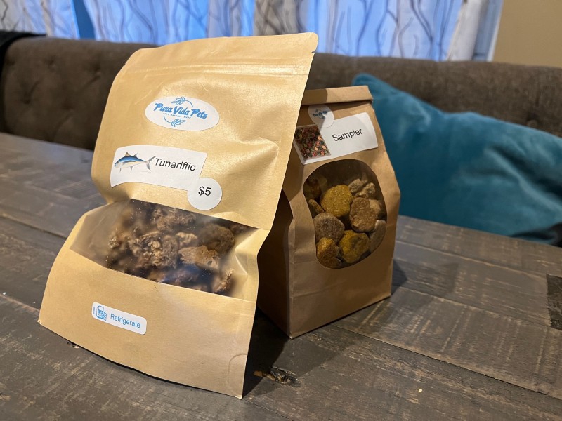 Two brown sacks of treats with clear windows are sitting on a gray wooden table. A taller bag has a sticker that says Tunariffic, and there are brown morsels visible. A shorter bag has a sticker that says Sampler, and there are small round cookie-like treats visible. Photo by Julie McClure.