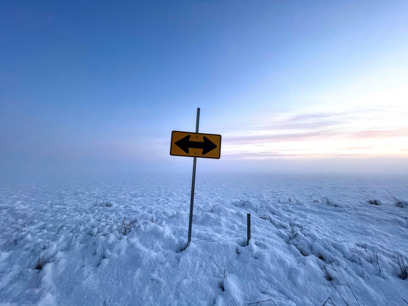 A wide snowy landscape with a bright blue sky. In the foreground is a sign post with a yellow sign with a black arrow pointing in either direction. Photo by Andrew Pritchard.