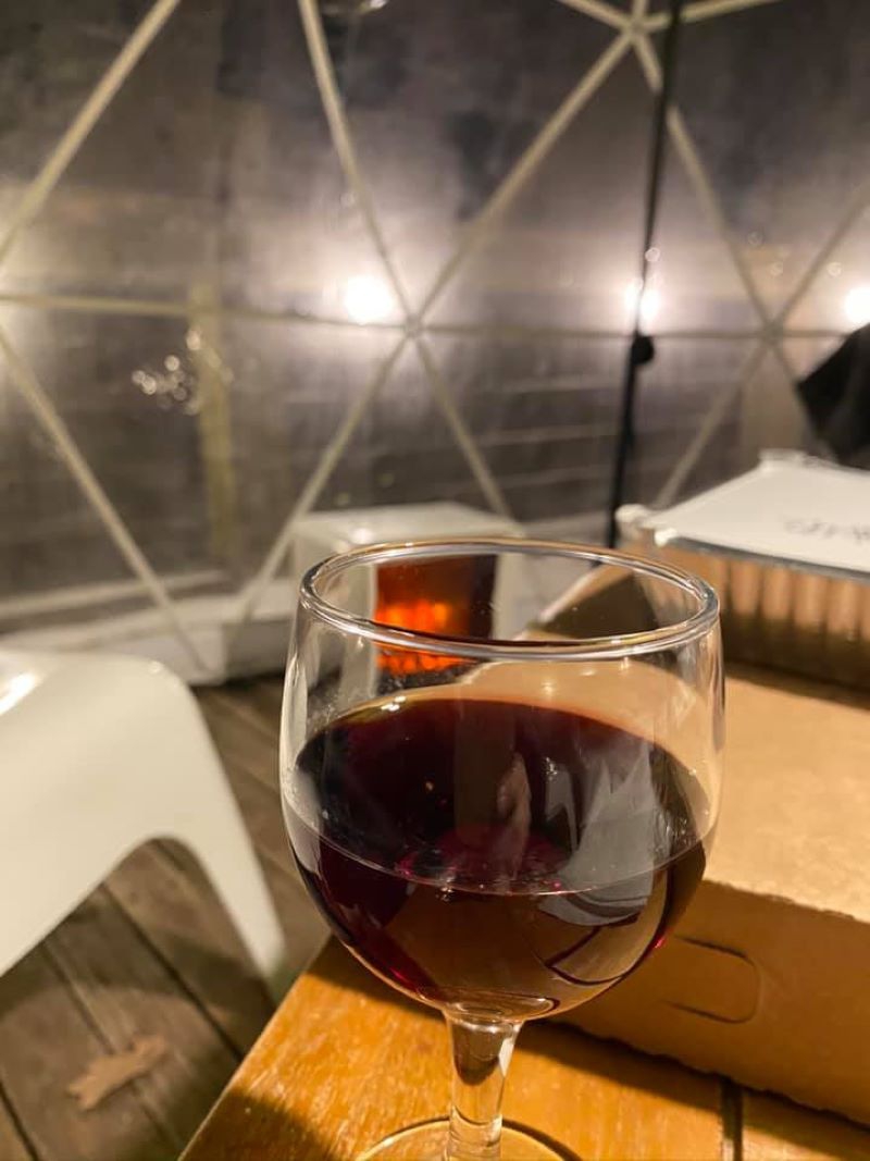 A glass of red wine is sitting on a wooden table. There is a pizza box behind the glass. In the background are the clear igloo-like walls of the dome. Photo by Julie McClure.