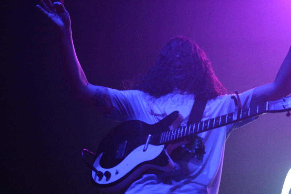 This is a photo of a musician playing a guitar on stage. The musician is wearing a white t-shirt and has long curly hair. The guitar is black and white and the musician is holding it up in the air. The background is dark with purple lighting. The musician’s arms are outstretched and they appear to be in the middle of a performance.