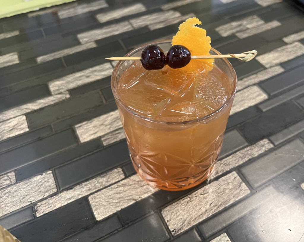 A rocks glass with an orange liquid, square ice, and a toothpick with two dark cherries.