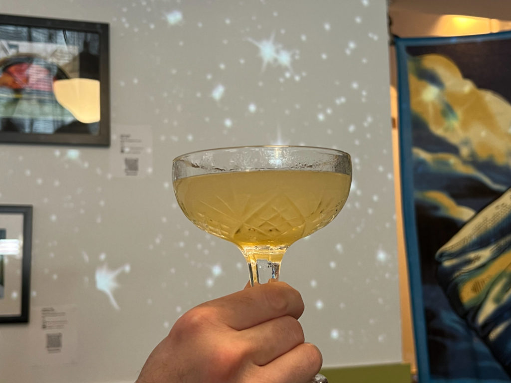 A coupe glass with a golden yellow drink in front of a white wall with projected starbursts.