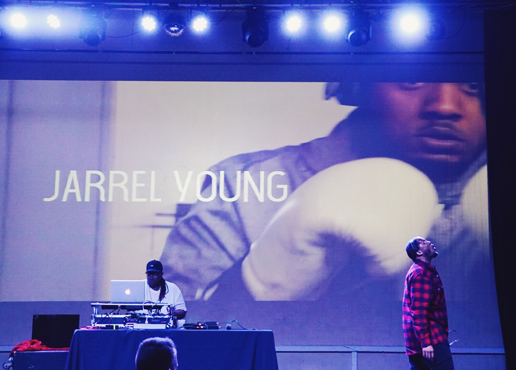 A man stands on stage, wearing a red and black checkered shirt, with a DJ behind him and a large screen displaying the name "JARREL YOUNG" along with an image of a man wearing boxing gloves.