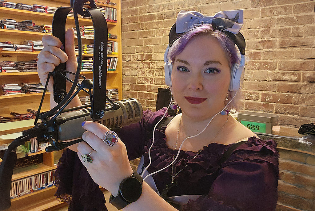 A woman is seated in a cozy studio filled with CDs and holding a professional microphone in her hand. She is wearing a dark purple dress with a matching headband adorned with a bow, and large white headphones. Her accessories include a colorful ring and a smartwatch.