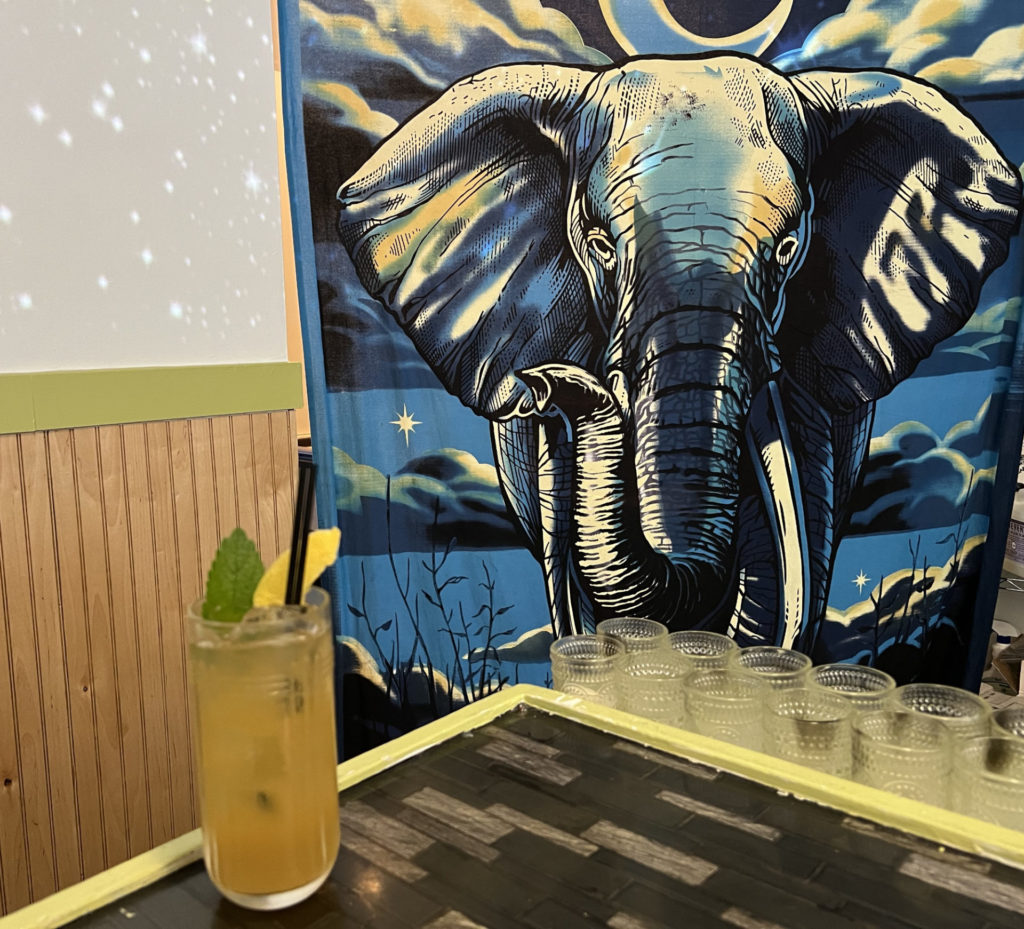 A tall glass with a yellow liquid ona counter that has a blue elephant tapestry behind it.