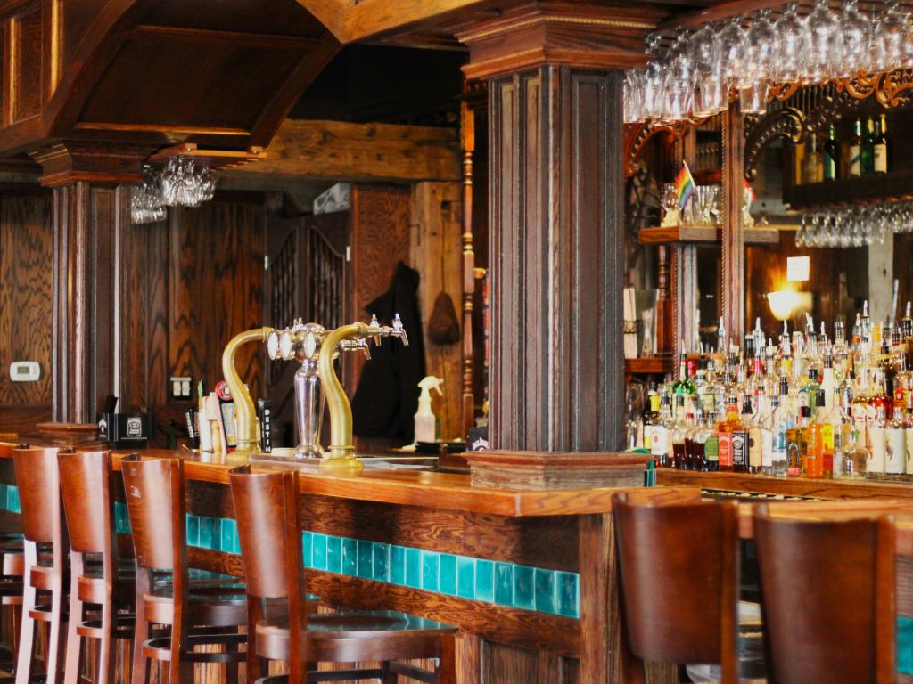 an interior of a wooden bar top with soft lighting and a bar full of bottles in front of a large mirror. The barstools are chairs with leather wrapped backs.