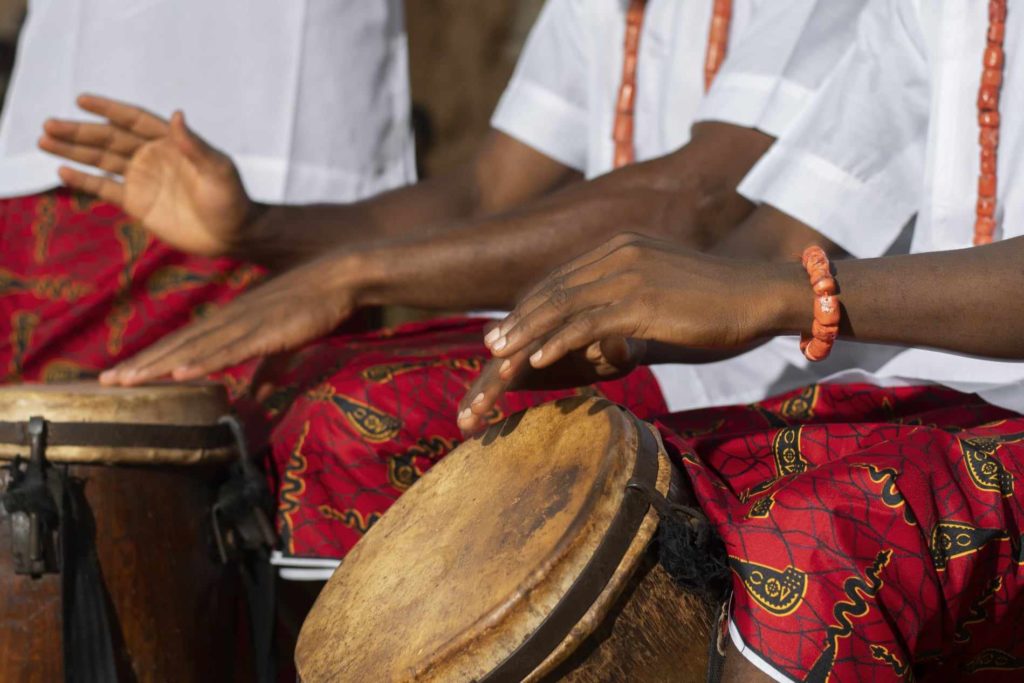 Musicians playing bongo drums wearing white shirts and textured red pants, orange necklaces and bracelets