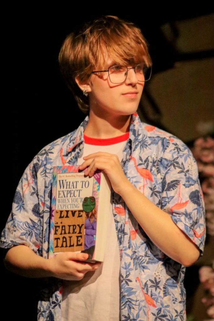 A young white person with glasses holds a book on a stage.