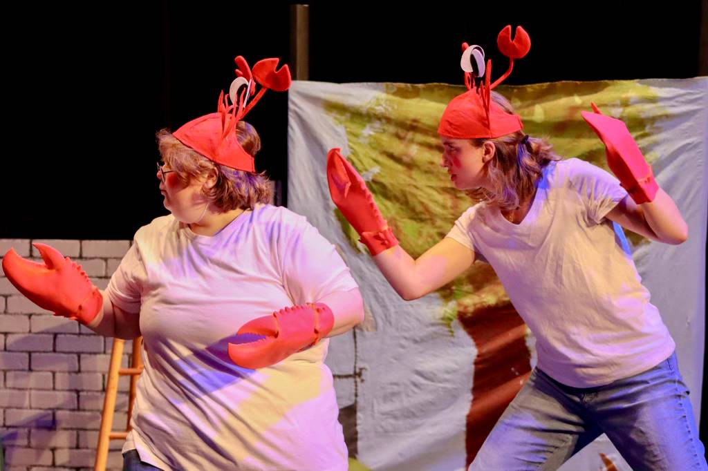 Two young actors are on stage in tee shirts and jeans and have portions of a red lobster hat and claws.