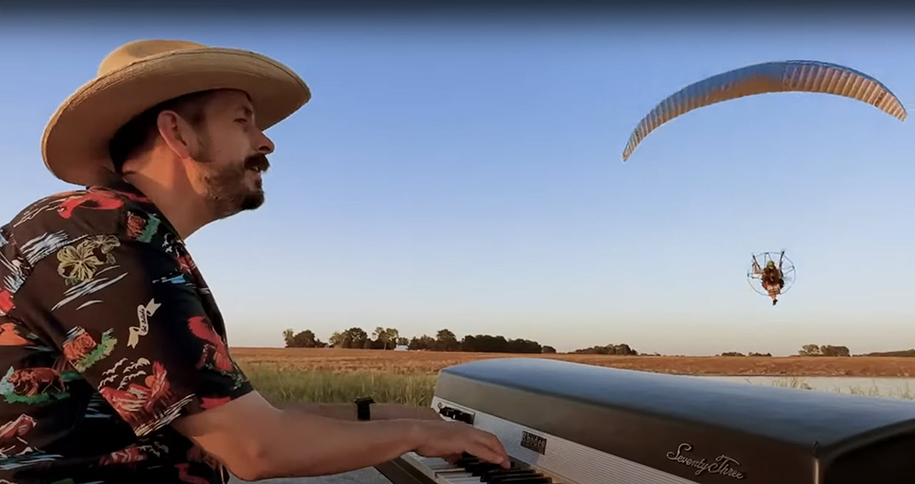 A man with a hat on plays an organ outdoors in a field with a paraglider whooshing by him already in the background.