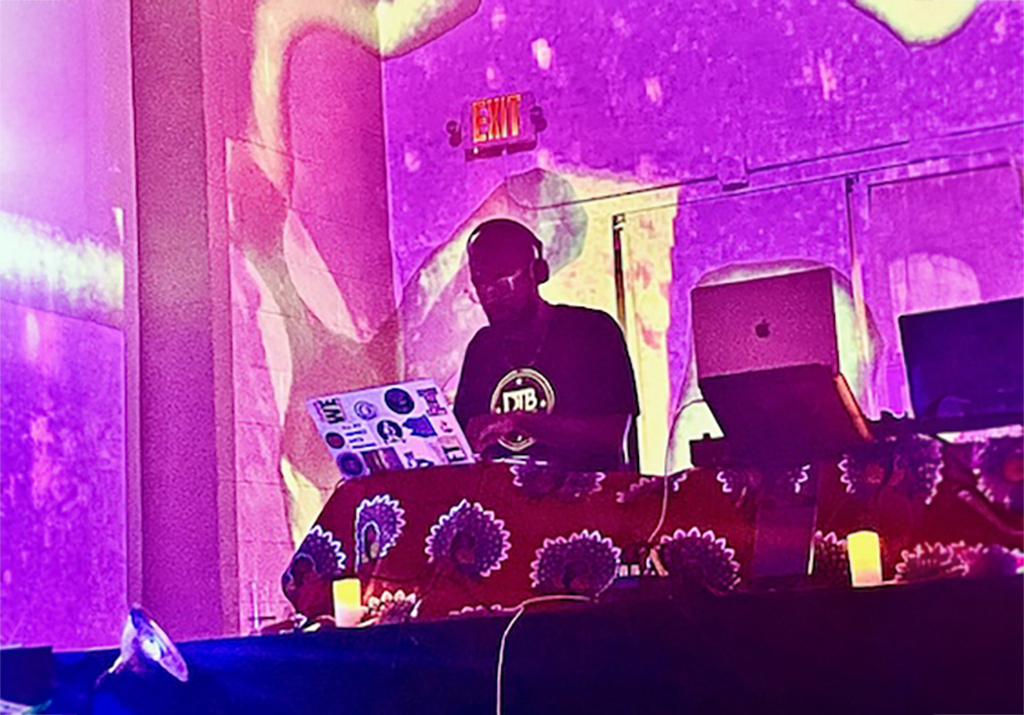 A DJ stands at a table with two laptops and various DJ equipment. The DJ is in a room with purple and green abstract light projections on the walls, creating a vibrant and immersive atmosphere. Several pieces of framed artwork are displayed on the walls, complementing the colorful projections. The room has an industrial feel with exposed ductwork on the ceiling. The overall scene suggests a lively and artistic event, blending visual art with music.