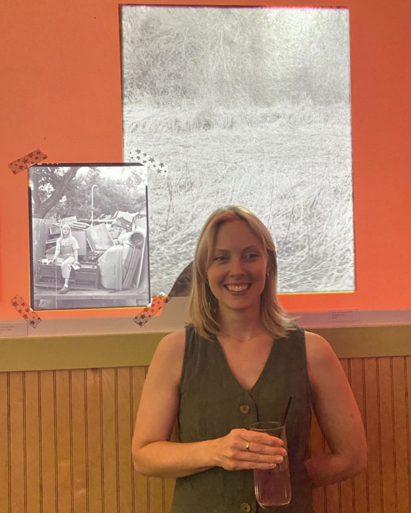 Photo of a white woman holding a drink in her right hand. She stands in front of photographs of herself in the middle of grey trash heap and a fuzzy image of prairie.