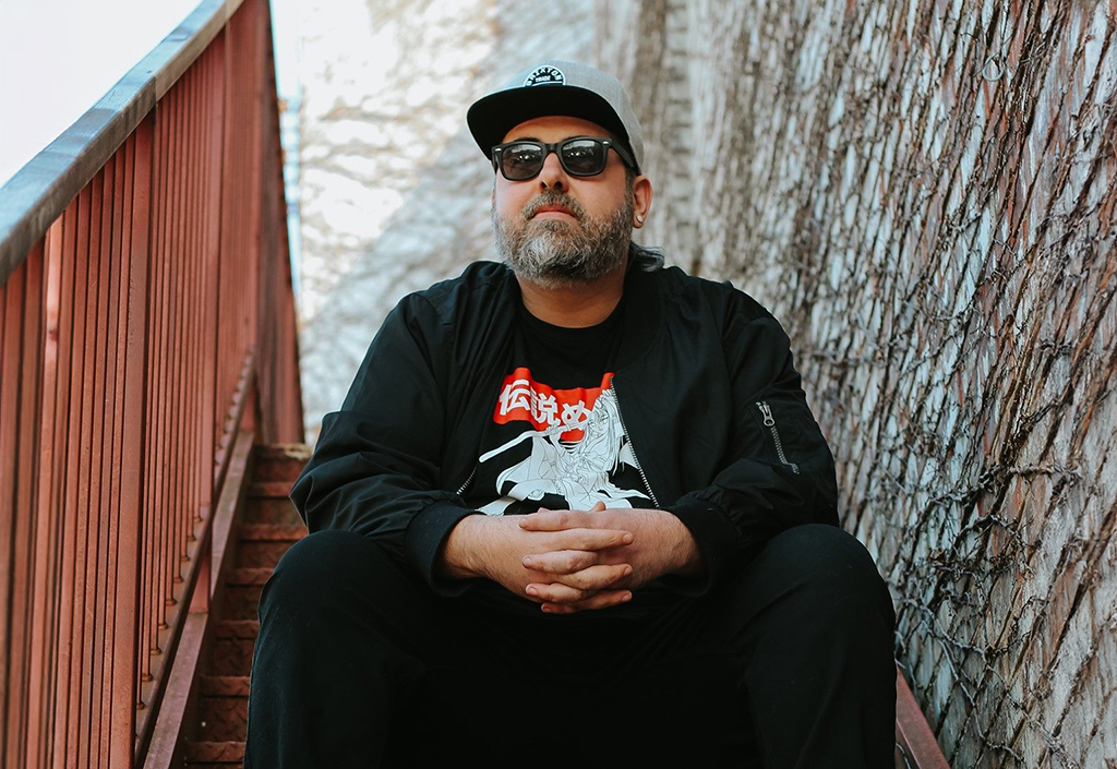 A man dressed in black with red and white artwork on his shirt sits on some stairs. To his right is a railing and to his left is a brick wall with Ivy on it. He has a grey hat and sunglasses on, and a salt and pepper beard.