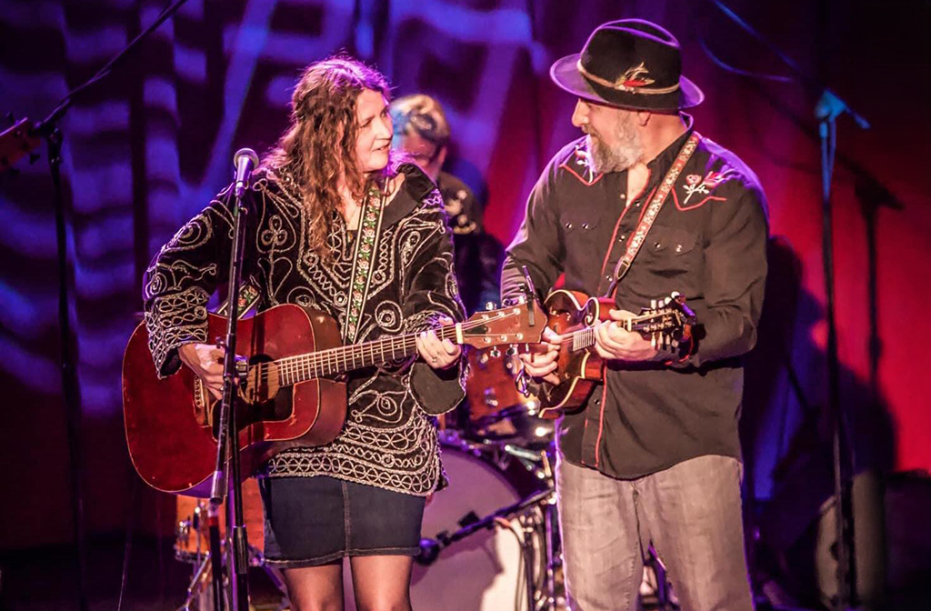 A duo, comprising a woman playing an acoustic guitar and a man playing a mandolin, is performing on stage. Both are wearing patterned outfits, with the woman in a detailed black dress and the man in a hat and black shirt, against a backdrop of purple lighting.
