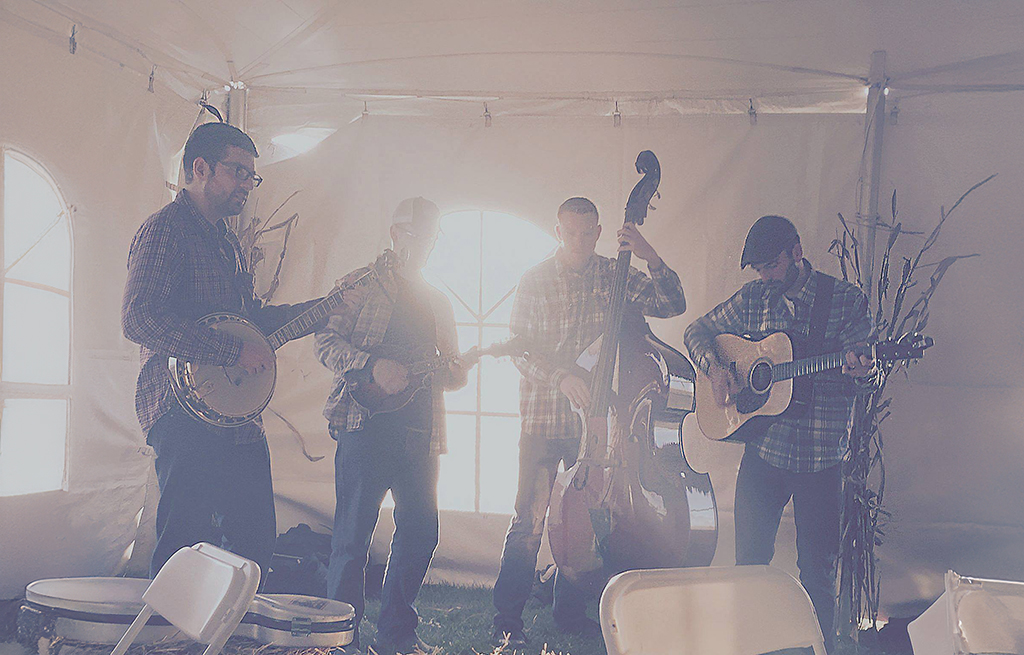 A bluegrass band performing inside a tent. The four members are playing banjo, mandolin, double bass, and acoustic guitar. They are all dressed in plaid shirts and hats, standing in a line with the natural light filtering through the tent's windows behind them. The setup is simple and rustic, with a casual and intimate feel to the performance.