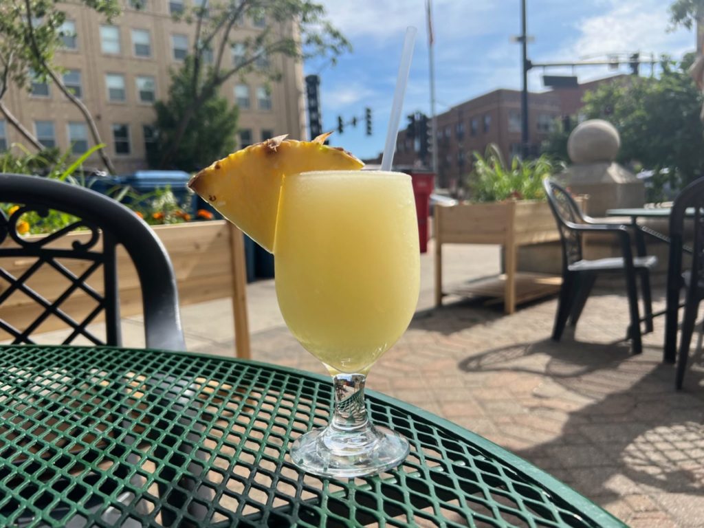 A glass goblet full of pineapple slush with a slice of pineapple and a clear plastic straw on a patio table in Downtown Champaign.