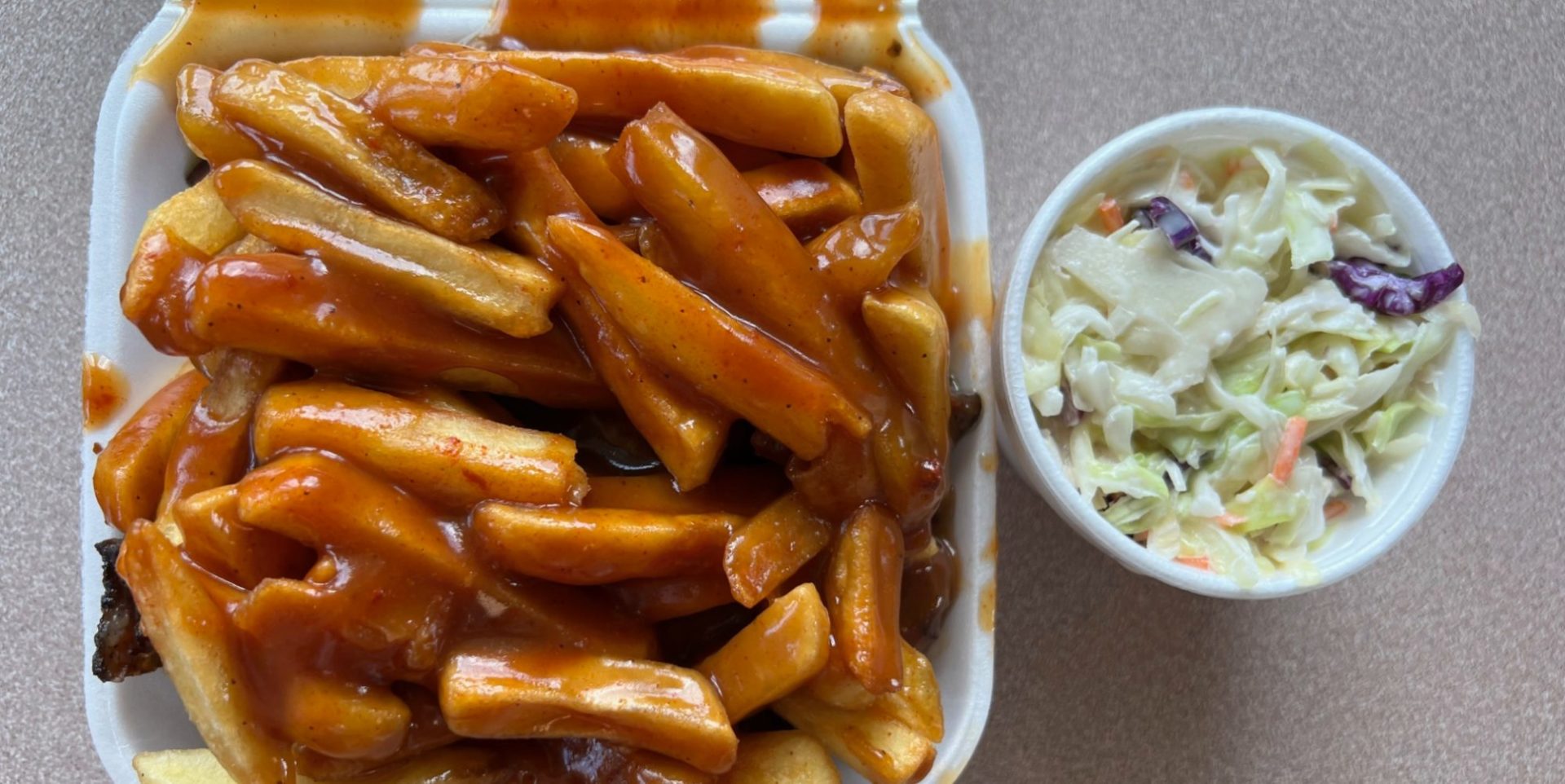A styrofoam container of jerk rib tips topped with fries and barbecue sauce beside a styrofoam cup of coleslaw.