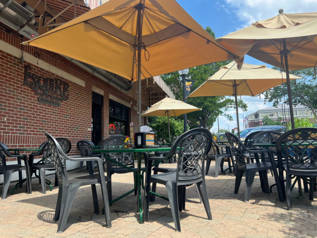 The outdoor patio of Esquire Lounge has black plastic patio chairs tucked into green metal tables with beige matching umbrellas.