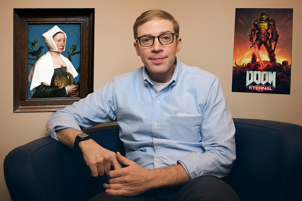 A man with short blond hair and glasses is sitting on a dark blue couch. He is wearing a light blue button-up shirt and brown pants, with a watch on his left wrist. Behind him on the wall, there is a framed painting of a woman in a white headscarf holding a fruit and a poster for the video game "Doom Eternal" featuring a muscular character with weapons.