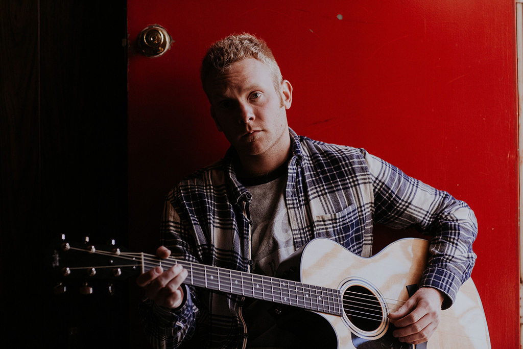 A color photograph of a man sitting in front of a bright red door, playing an acoustic guitar. He is wearing a plaid shirt over a t-shirt and has a serious, thoughtful expression. The lighting is warm, creating a cozy and intimate setting.