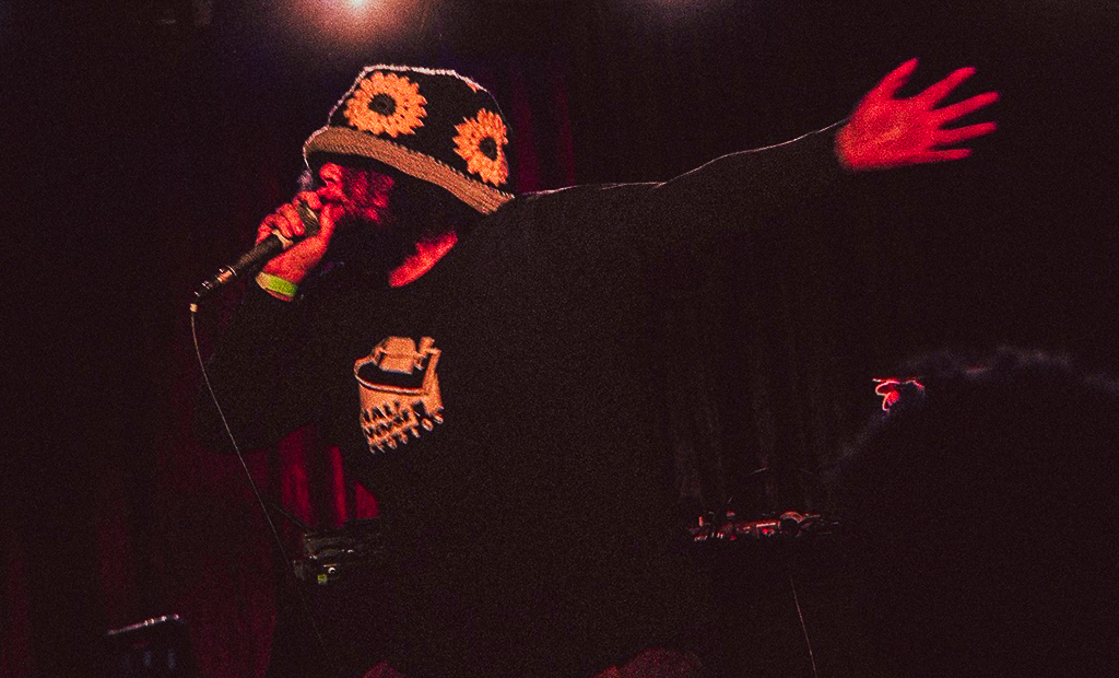 A performer is on stage, captured mid-performance. He is wearing a black shirt with a graphic of a typewriter and a colorful crocheted bucket hat with large sunflower designs. He holds a microphone close to his mouth with one hand, while the other arm is extended outward, creating a dynamic and expressive pose. The background is dark, with stage lights casting a dramatic effect, highlighting the intensity of the performance.
