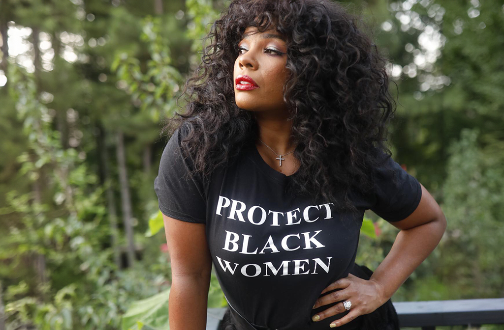A woman stands outdoors in front of a backdrop of green foliage. She has voluminous, curly hair cascading around her shoulders and wears a black T-shirt with white lettering that reads "PROTECT BLACK WOMEN." She also sports bright red lipstick and a cross necklace, adding to her striking appearance. One hand rests confidently on her hip, highlighting a large ring on her finger. The setting appears to be a natural, wooded area, providing a serene contrast to her bold and powerful presence.