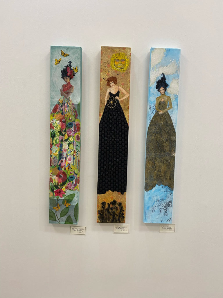 Photo of three textured paintings of women with exaggerated hair and dresses in black, floral print, and green against blue sky, sunlight, and butterflies