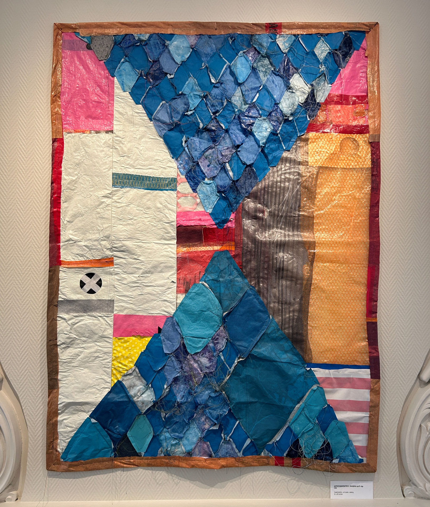 Brightly colored quilt made of plastics handing on a wall--bright blues and pink shapes make up the bulk of this quilt.