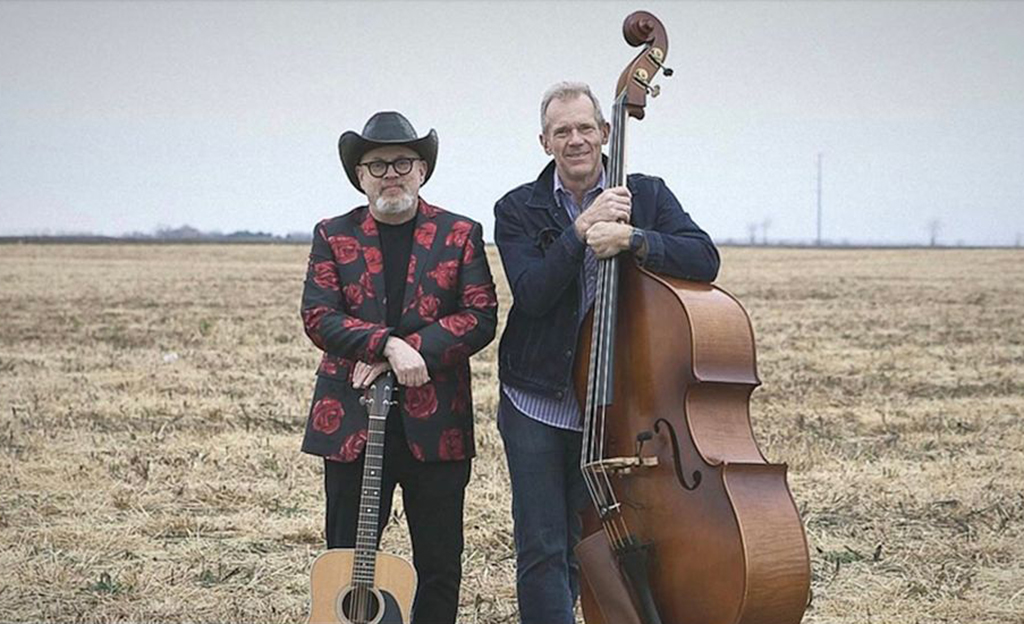 Two musicians stand in the field with their instruments. The person on the left is wearing a black cowboy hat and has a guitar. The person on the right has an upright base.
